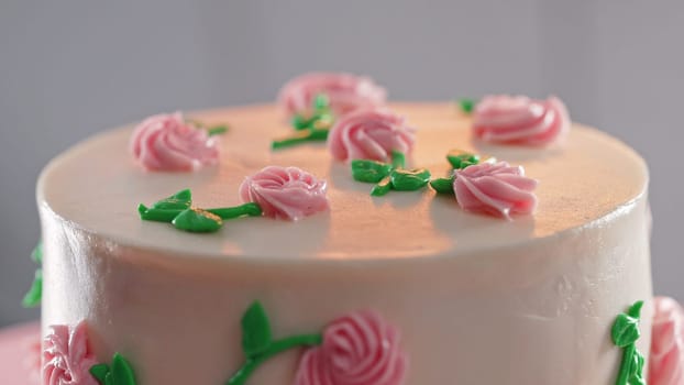 Beautiful buttercream cake on turntable. Decorated with delicate pink flowers, green leaves on smooth white icing. Bakery, celebration, wedding, pastry concept. High quality
