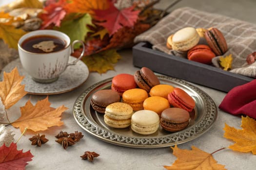 Assorted macarons in vibrant colors rest on a silver platter, accompanied by a warm cup of coffee. Autumn leaves surround the setting, enhancing the cozy atmosphere.