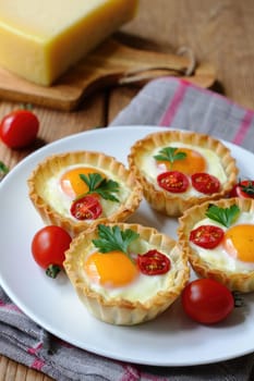 Exquisite tartlets filled with eggs, cherry tomatoes, and fresh herbs are displayed on a rustic wooden table. Cheese sits nearby, adding to the inviting atmosphere of this culinary delight.