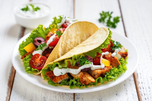 Two grilled chicken tacos filled with lettuce, served alongside a vibrant salad of tomatoes, peppers, and olives. A small bowl of creamy dressing completes the meal.