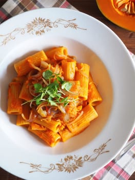 A colorful bowl of rigatoni pasta topped with savory pumpkin sauce and garnished with fresh herbs sits on a decorative table. The dish combines vibrant colors and textures.