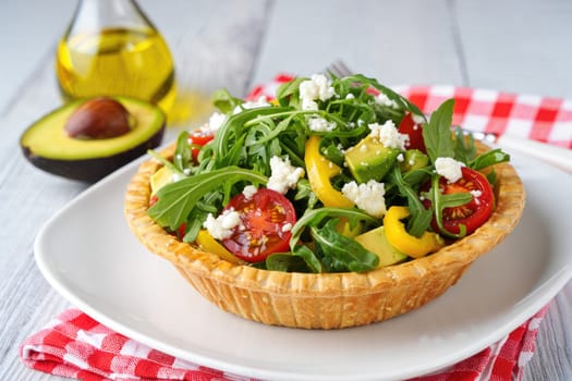 A colorful salad featuring mixed greens, cherry tomatoes, yellow peppers, and feta cheese is served in a golden pastry crust. Olive oil and an avocado accompany the dish on a rustic wooden table.
