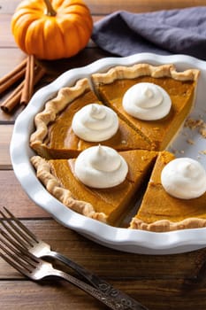 A freshly baked pumpkin pie with a golden crust and dollops of whipped cream sits on a wooden table. Cinnamon sticks and a small pumpkin accompany the pie, capturing the essence of autumn.