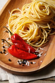 Twirls of pasta sit elegantly on a wooden plate, accompanied by two vibrant red chili peppers and a sprinkle of coarse salt and black pepper, creating a tempting presentation.