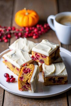 Savor the taste of freshly baked pumpkin cranberry bars topped with cream cheese frosting. The warm dessert is beautifully arranged on a plate beside a cup of coffee and autumn decorations.