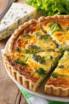 A delicious broccoli quiche sits on a wooden table, showcasing a golden crust and vibrant green broccoli. Nearby, a wedge of blue cheese adds a touch of gourmet flair, accompanied by fresh greens.