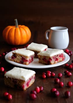 Soft cranberry pumpkin bars topped with cream cheese icing are displayed on a white plate. Fresh cranberries and a small pumpkin add to the cozy autumn ambiance of this dessert presentation.