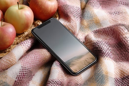Modern smartphone placed on a warm checkered wool blanket surrounded by apples. Ideal for autumn campaigns, tech branding, rustic lifestyle visuals, or cozy seasonal product presentations