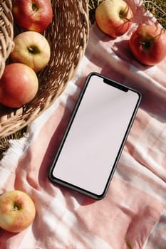 Smartphone lying on a red-and-white picnic blanket next to fresh apples and a wicker basket in natural sunlight. Ideal for lifestyle themes, product mockups, or autumn branding scenes