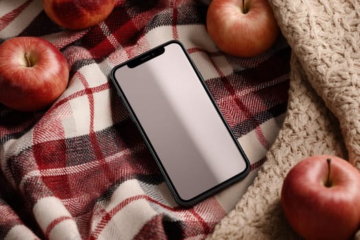 A smartphone on a red plaid blanket with apples and knitted fabric in warm, soft lighting. Great for autumn-themed promotions, lifestyle scenes, and mobile app mockups