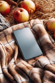 A smartphone placed on a soft brown plaid blanket with fresh apples and rustic hay in the background. Ideal for autumn tech ads, cozy lifestyle visuals, and seasonal promotions