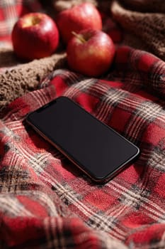 Smartphone placed on a soft red plaid blanket next to fresh apples and rustic knits. A cozy autumn scene perfect for seasonal tech ads, lifestyle content, and fall marketing visuals