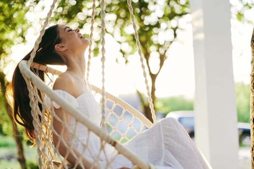 A joyful young woman in a flowing white dress sits on a swinging chair, surrounded by lush greenery and sunlight, embodying peace, happiness, and relaxation in nature