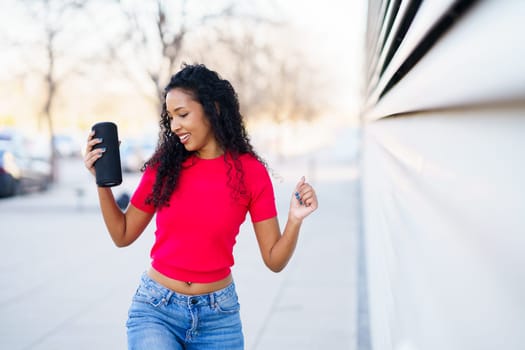 A joyful woman with curly hair dances exuberantly in an urban setting, holding a drink. Wearing a stylish red top and trendy jeans, she radiates fun and positivity, celebrating happiness
