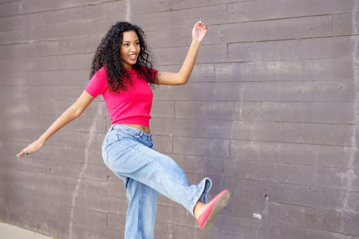 A vibrant young woman dressed in a cheerful pink top and trendy baggy jeans poses joyfully against a textured gray wall, showcasing her playful style, infectious energy, and colorful personality
