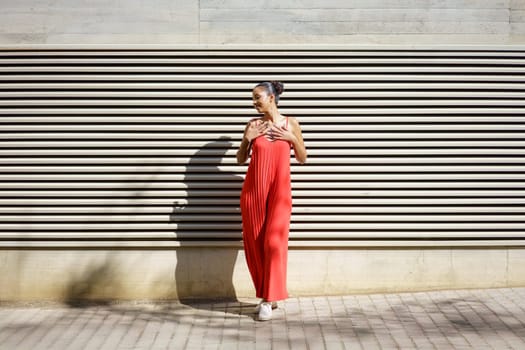 A stylish and fashionable woman poses confidently in a vibrant and eyecatching red dress, standing proudly against a modern textured wall, showcasing her contemporary style and elegant personality