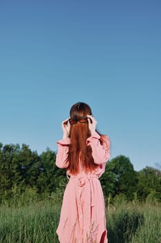 A woman wearing a pink dress stands in a green field, playfully obscuring her face with her long hair The sunny blue sky enhances the feeling of freedom and joy in outdoor fashion