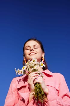 Happy young woman in a pink shirt embraces nature while holding wildflowers under a vibrant blue sky, embodying joy, and tranquility