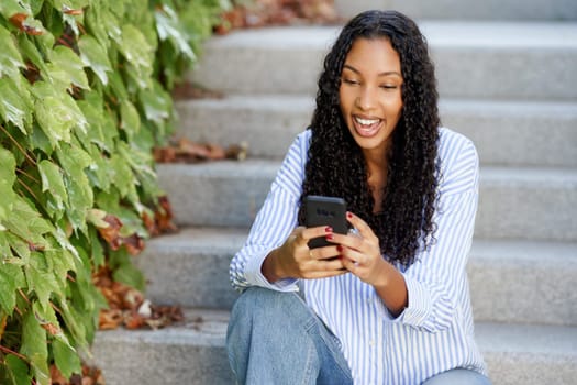 A cheerful young woman with curly hair is seated on steps, smiling as she uses her smartphone. Lush green ivy frames the scene, creating a vibrant atmosphere of warmth and happiness