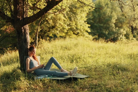 A serene young woman in comfortable fitness attire reclining against a tree, embodying relaxation and wellness in a lush green setting, ideal for promoting a healthy lifestyle or outdoor activities