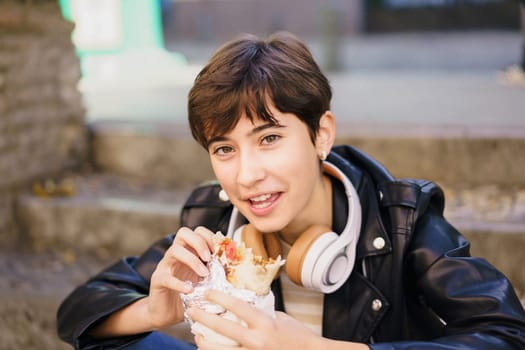 In a lively urban setting filled with energy and movement, a young person sporting a trendy short hairstyle and wearing headphones is happily sitting on the stairs while enjoying a delicious burrito,