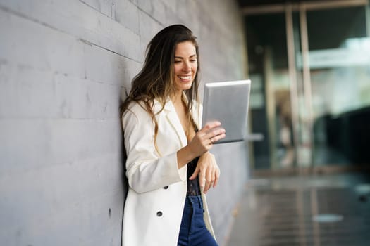 A young woman smiles brightly as she uses her tablet, dressed stylishly while leaning against a wall in an urban setting, showcasing her relaxed demeanor and enjoyment of the digital interaction