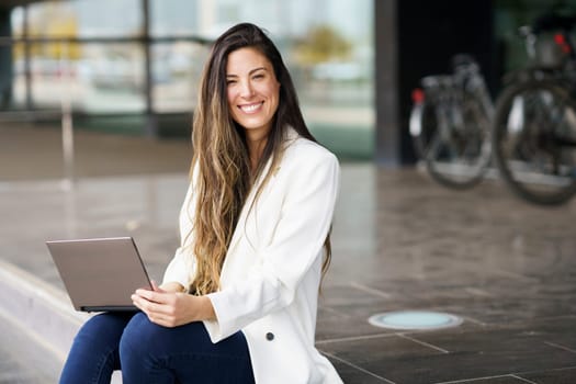 A cheerful young woman with long flowing hair beams with joy as she engages with her laptop outdoors in a vibrant modern setting, showcasing a perfect blend of leisure, technology, and nature