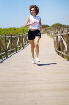 Full body of active female athlete in sportswear and sneakers running along wooden boardwalk during fitness training