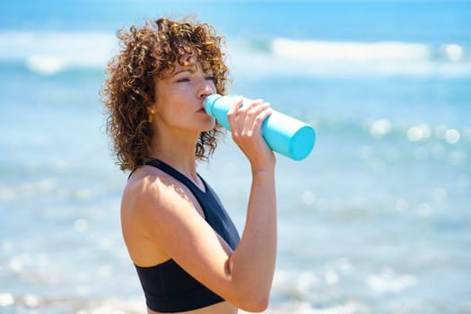 Side view of young fit sportswoman with curly hair drinking fresh water after training near waving sea on sunny day