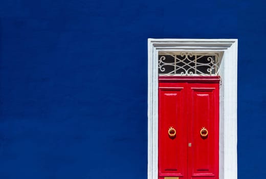 High contrast photograph of dark blue wall of residential building facade with red door in white wooden frame