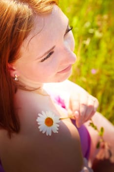 A woman with red hair sits in a lush green meadow, appreciating nature and gently holding a daisy against her skin, basking in sunlight.