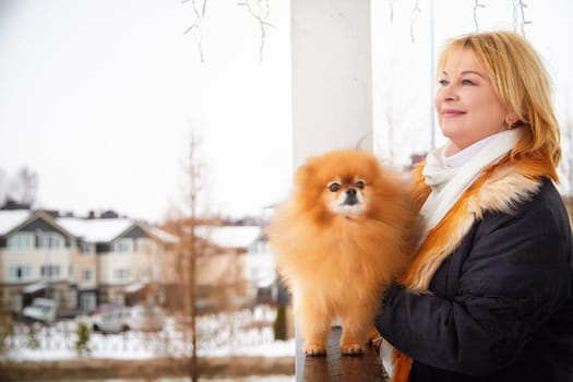 A woman holds her fluffy Pomeranian dog, enjoying a winter day in a snow-covered neighborhood with warm attire.