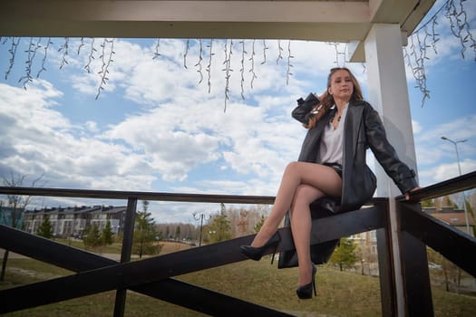 A young woman showcases her fashion while sitting elegantly on a balcony under a blue sky filled with clouds.