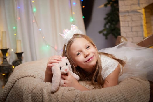 Girl in a beautiful holiday dress holds a plush bunny while enjoying the cozy ambiance of Christmas lights and decorations.