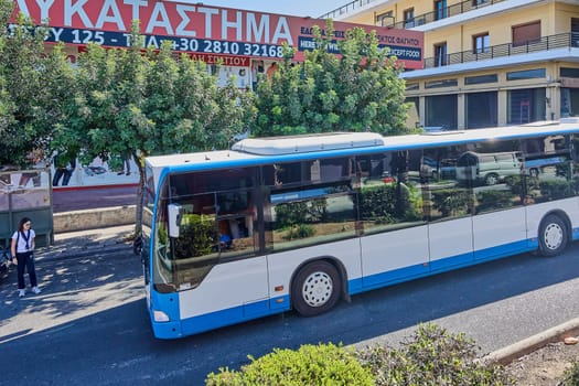 Crete.Greece - april 13, 2025: An urban bus in Heraklion, painted blue and white, parked near a bus stop. The bus reflects surrounding trees and vehicles, while a nearby sign advertises local food businesses.