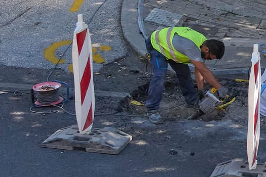 Crete.Greece - april 13, 2025: A hardworking laborer in Heraklion operates machinery during street maintenance. Tools and signs frame the active repair site under the clear Mediterranean sky.