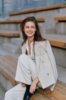 A confident woman dressed in a light-colored business outfit is seated outdoors, radiating positivity with a warm smile, set against a modern urban backdrop.