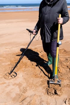 Elderly adult using metal detector on beach wearing black coat and boots.