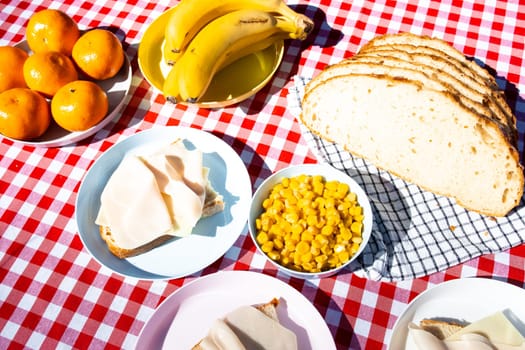 Outdoor picnic table with sliced bread, cheese, fruit, and corn on red checkered cloth.