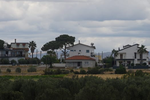 Nestled in a tranquil coastal village, quaint houses stand under an overcast sky, surrounded by lush greenery and distant hills, embodying a peaceful, serene atmosphere.