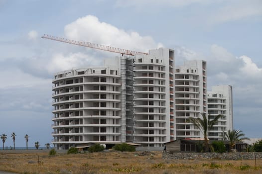 Amidst a tranquil landscape, a modern residential structure rises towards the sky, partially completed. The coastline glimmers in the background under a moody, cloudy sky.
