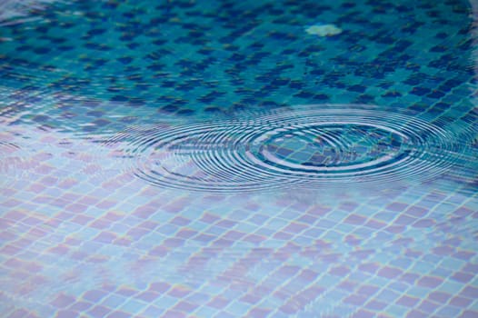 Gentle ripples expand across the shimmering surface of a serene pool, reflecting the sunlight and showcasing intricately arranged blue tiles. A peaceful moment unfolds in the warmth of the day.