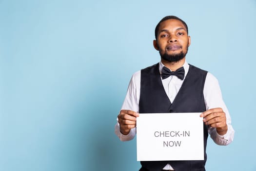 Professional doorman holding a paper sign for clients to go to room check in, presenting a message for attention. Hotel porter in a suit and tie standing against blue background.