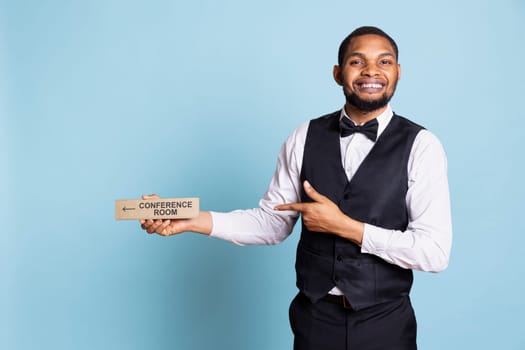 Front desk staff helping clients with indicators to all amenities in studio, giving directions and showing wall sign pointer for the conference room. Skilled doorkeeper in tuxedo.