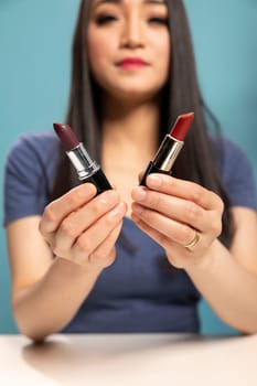 Elegant woman holding different lipstick shades, presenting them to the camera during her live broadcast session. Closeup of cosmetics items highlighted by asian beauty content creator.