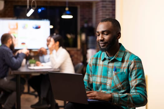 African american entrepreneur reviews financial planning data on device as startup team collaborates in background. Portrait of businessman with laptop, checking emails and planning his schedule.