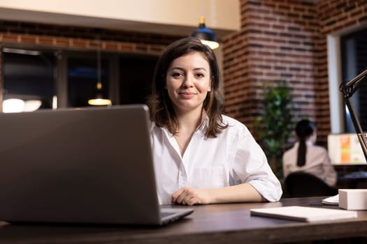 Businesswoman sitting at desk with her personal computer, in preparation to work a night shift in modern office. Portrait of female entrepreneur looking at camera, portraying confidence at workplace.