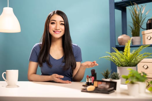 Cheerful beauty influencer presenting a makeup tutorial, creating content for her vlogging channel. Asian woman gestures with her hand while explaining the purpose of her video during live broadcast.