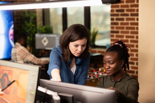 Multiethnic women closely collaborate on a digital art project in a marketing agency, reviewing designs on computer. Caucasian manager advising black female editor in modern workspace.