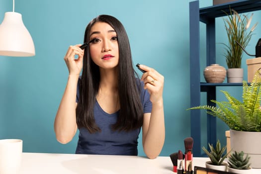 Beautiful woman using cosmetic product while recording a makeup tutorial video for online audience. Asian female vlogger seated at desk, demonstrating how to apply mascara during her live broadcast.